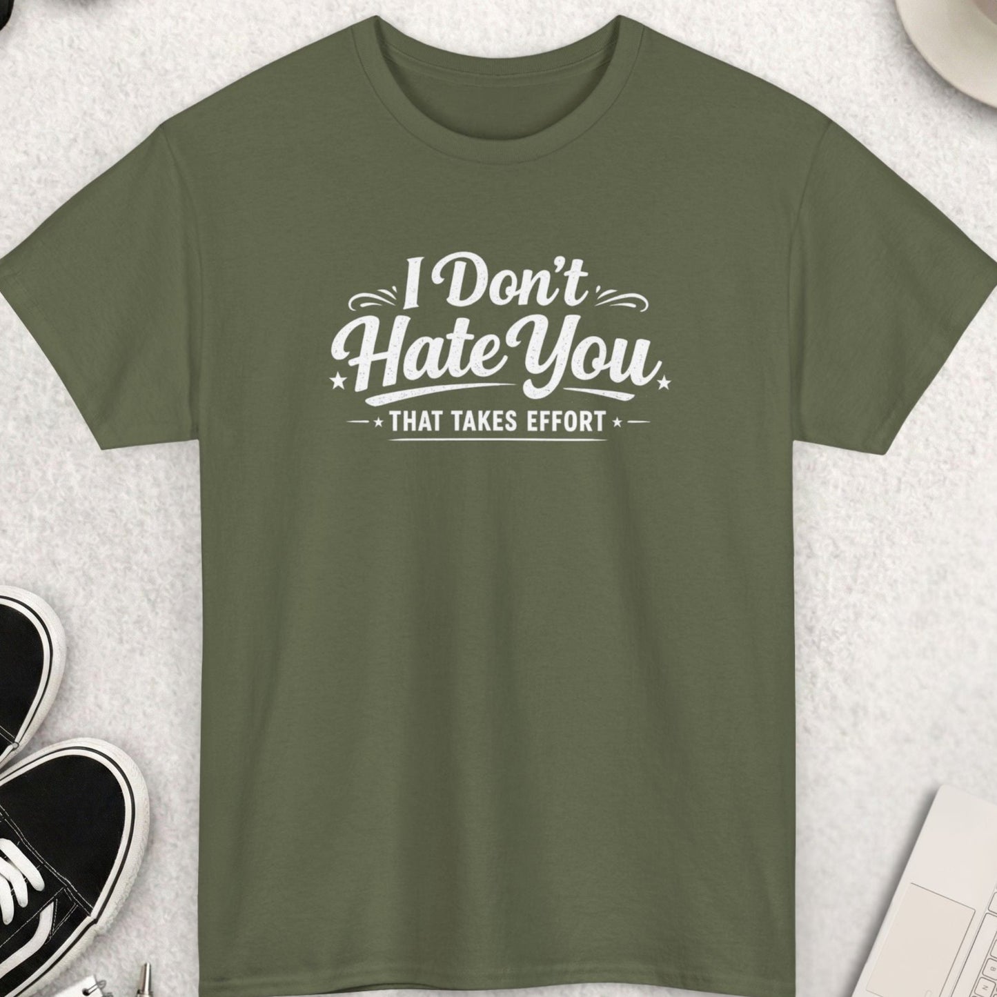 Green t-shirt with 'I Don't Hate You That Takes Effort' text on a light gray surface with shoes, coffee, and sunglasses.
