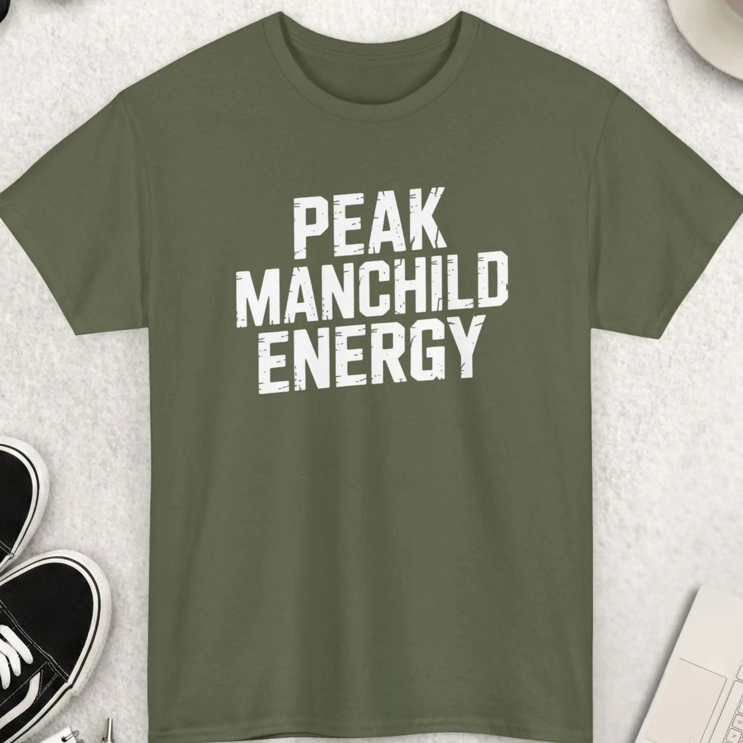 Green t-shirt with 'Peak Manchild Energy' text on a light gray surface with accessories.
