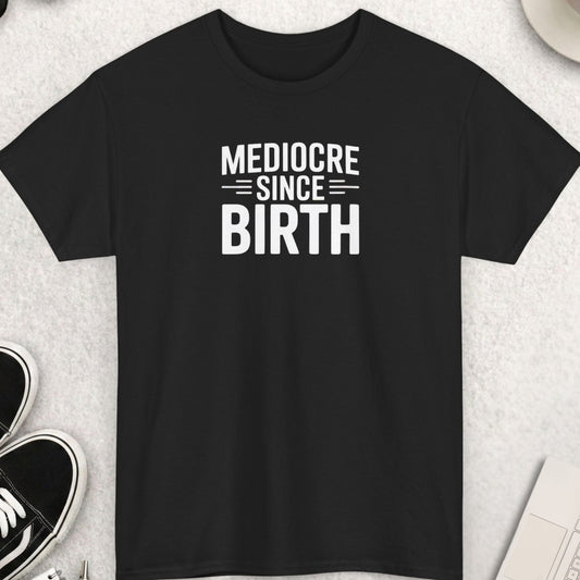 Black t-shirt with 'Mediocre Since Birth' text on a light gray surface with shoes, coffee, and sunglasses.