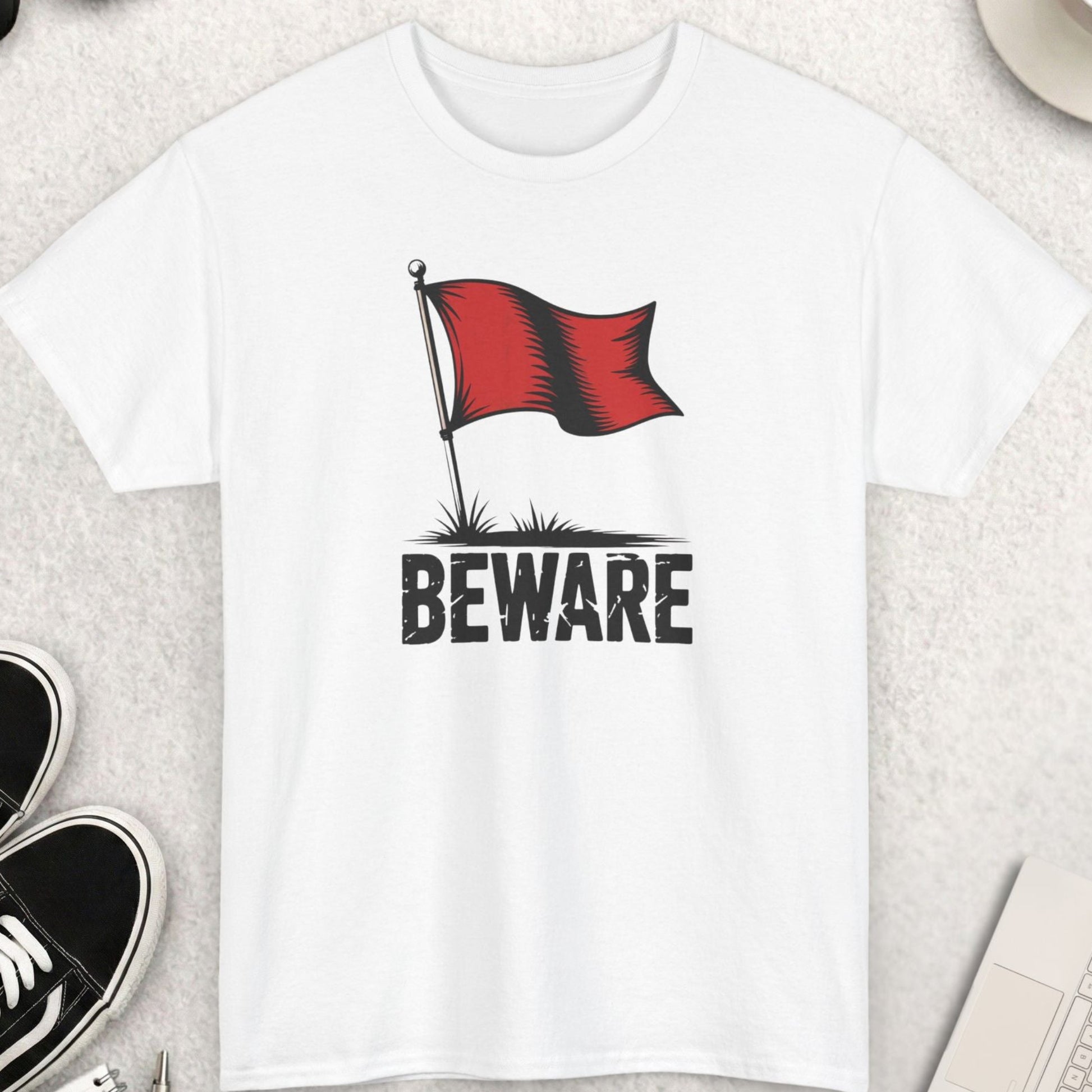 White t-shirt with a red flag and 'Beware' text on a light gray surface with shoes, coffee, and sunglasses.