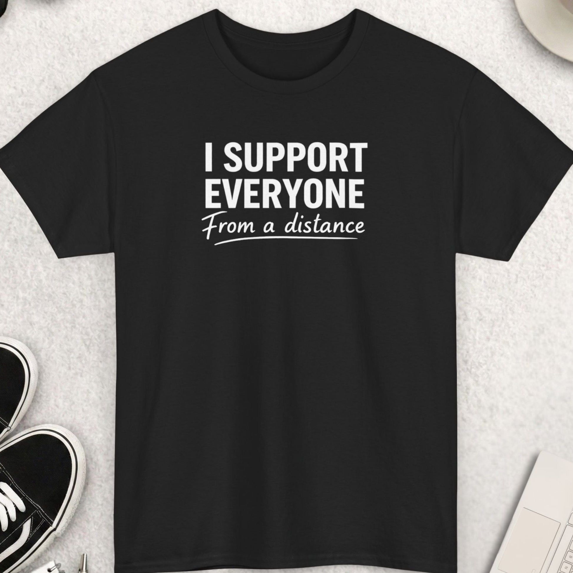 Black t-shirt with 'I Support Everyone From a Distance' text on a light gray surface with shoes, coffee, and accessories.