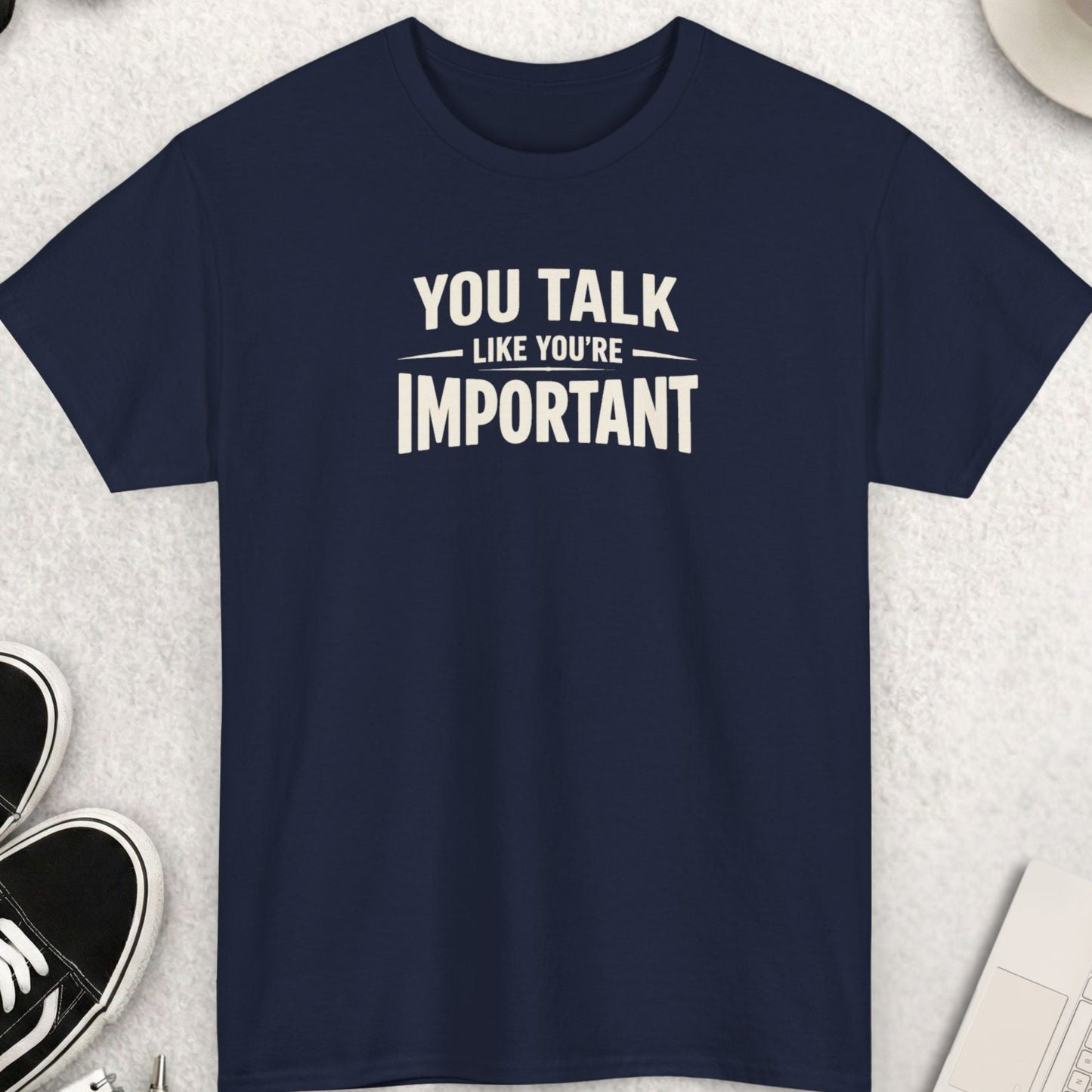 Navy blue t-shirt with 'You talk like you're important' text on a light gray surface with shoes, coffee, and accessories.