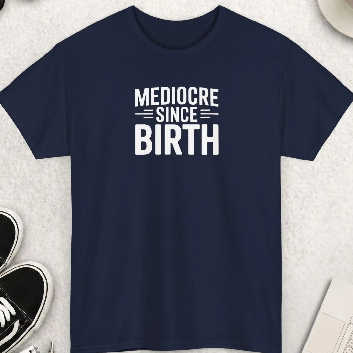 Navy blue t-shirt with 'Mediocre Since Birth' text on a light gray surface with accessories.