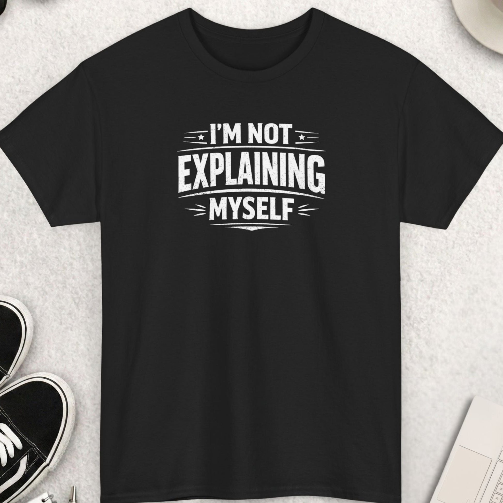 Black t-shirt with 'I'm not explaining myself' text on a light gray surface with shoes, coffee, and accessories.