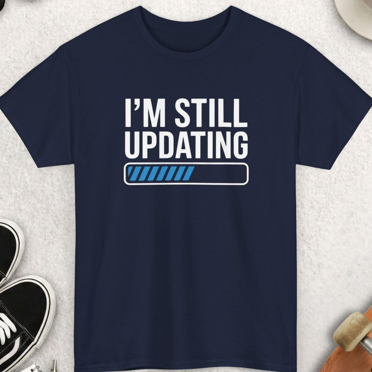 Navy blue t-shirt with 'I'm Still Updating' text on a light gray surface with accessories.