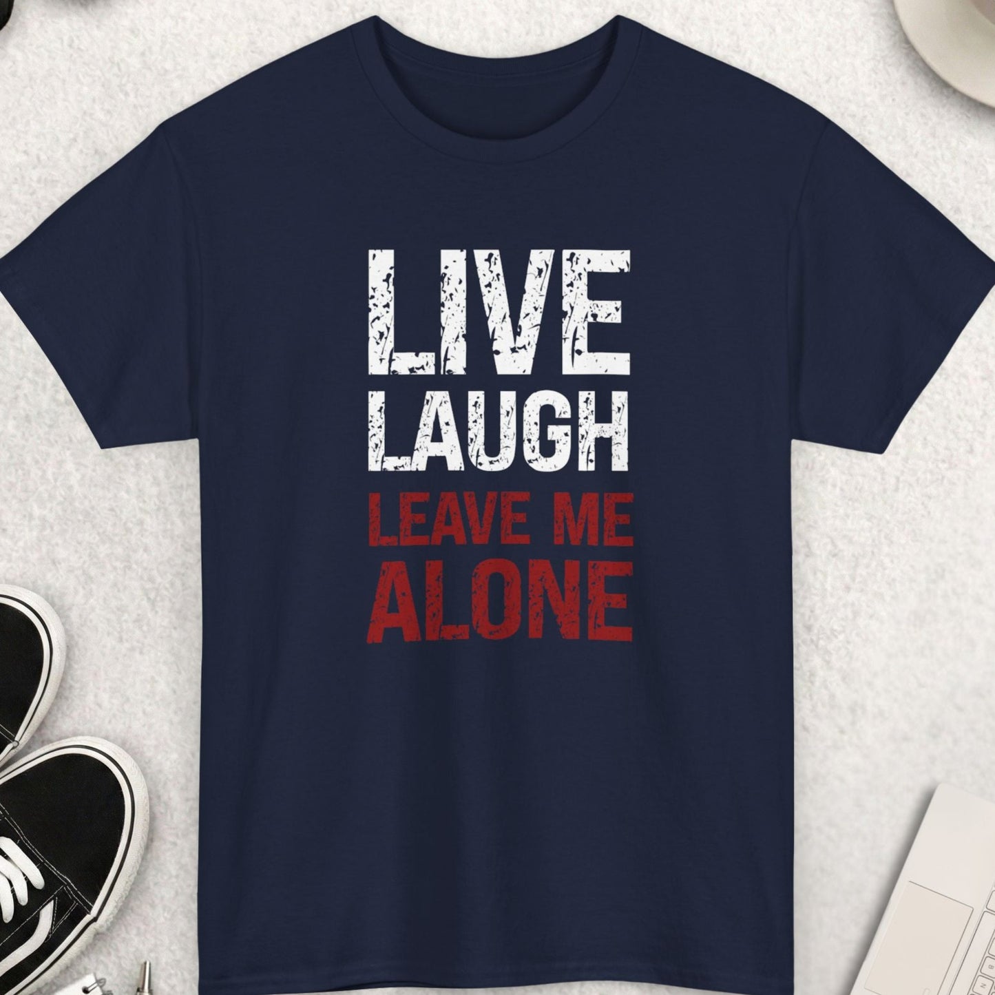 Navy blue t-shirt with 'Live Laugh Leave Me Alone' text on a light gray surface with accessories.