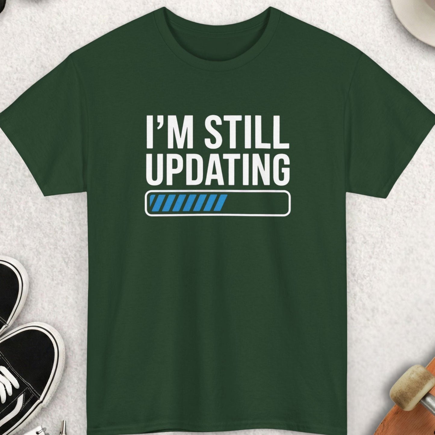 Green t-shirt with 'I'm Still Updating' text on a light gray surface with accessories.