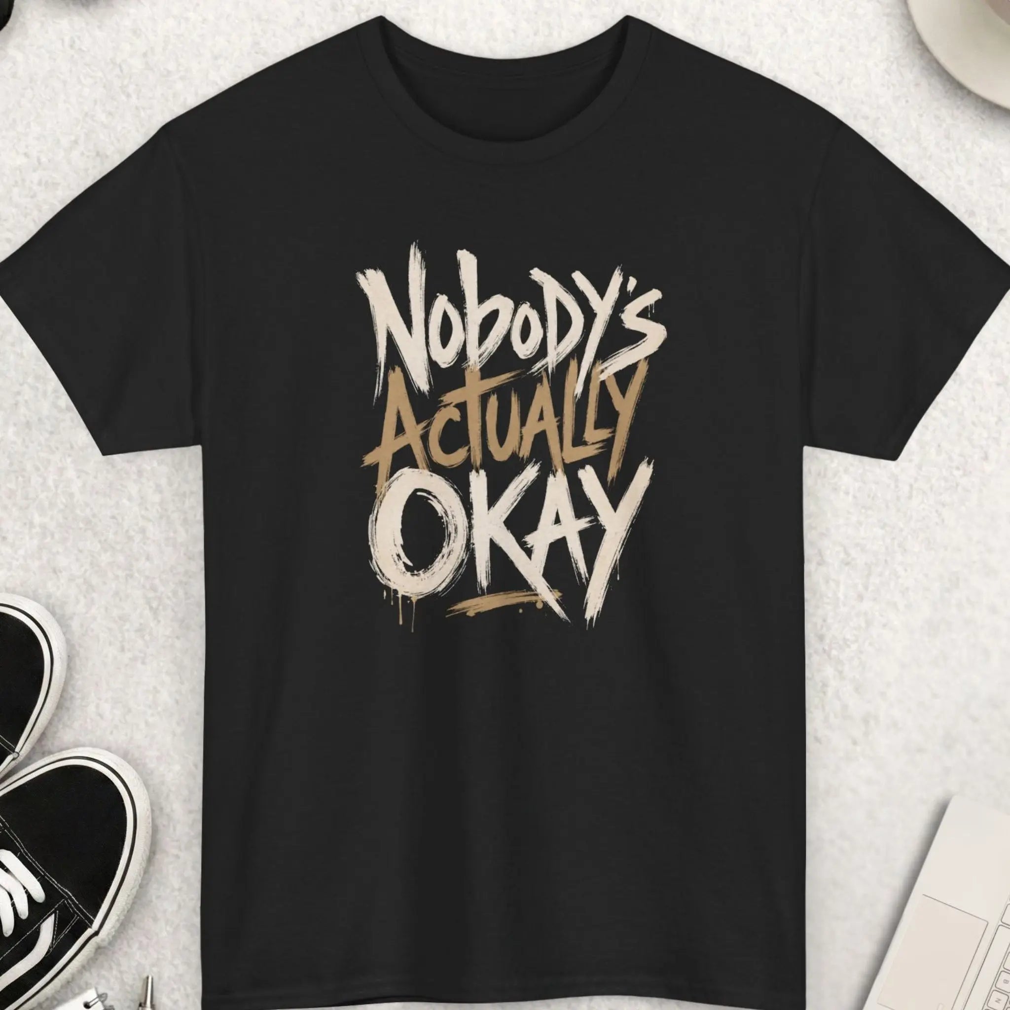 Black t-shirt featuring expressive distressed brush typography reading “Nobody’s Actually Okay” in a bold hand-painted minimalist design.