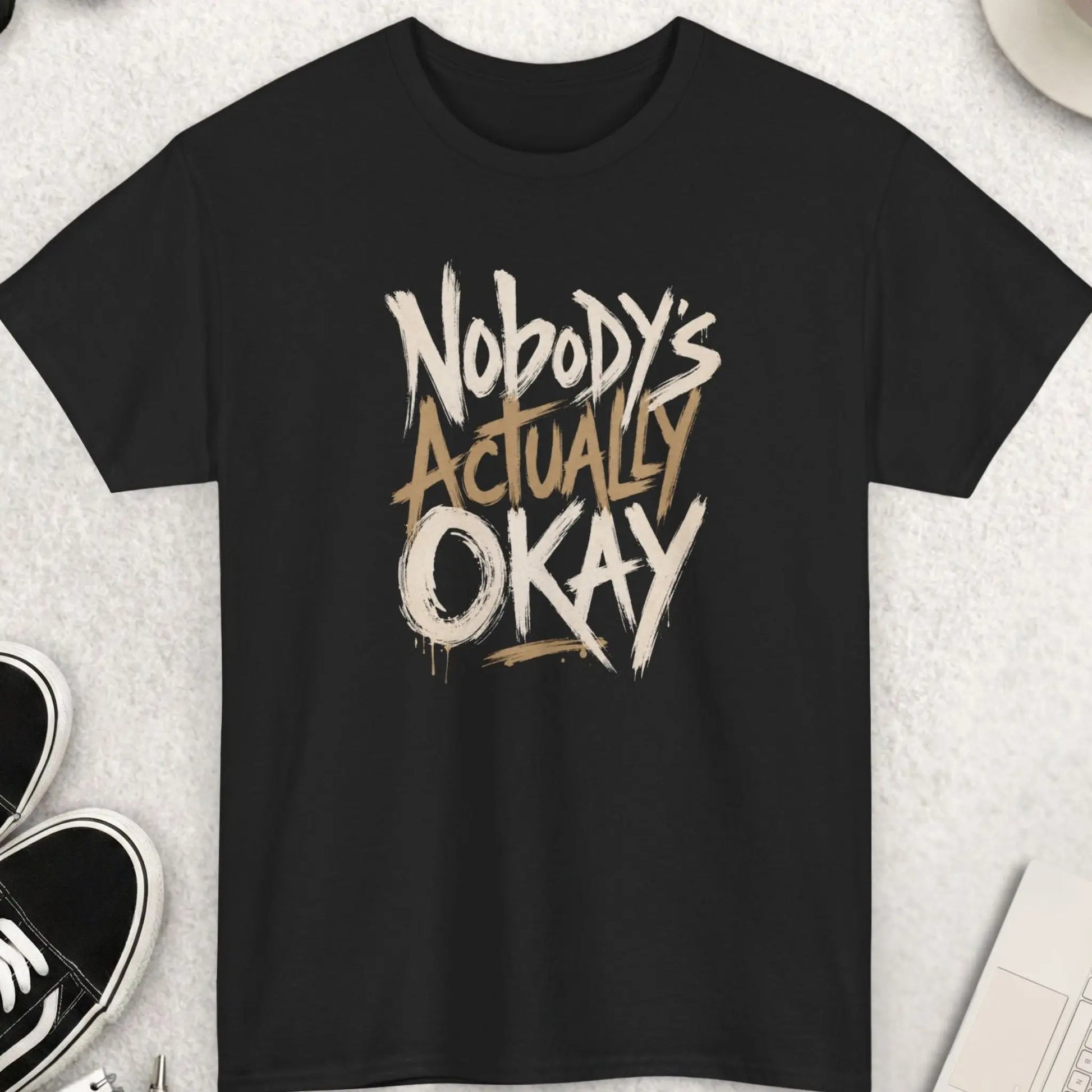 Black t-shirt featuring expressive distressed brush typography reading “Nobody’s Actually Okay” in a bold hand-painted minimalist design.