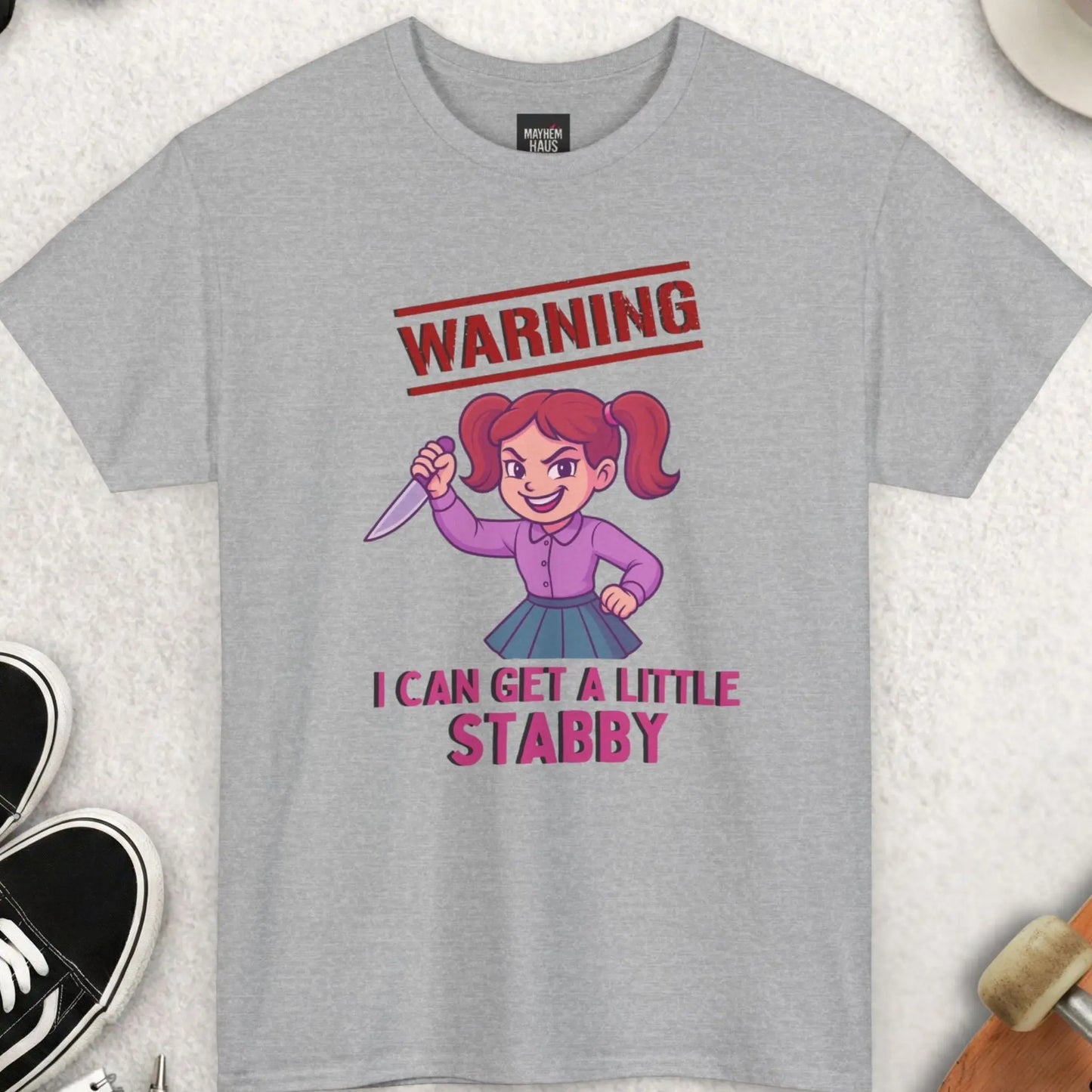 Gray t-shirt with cartoon character and text 'Warning: I can get a little stabby' on a textured surface with accessories.