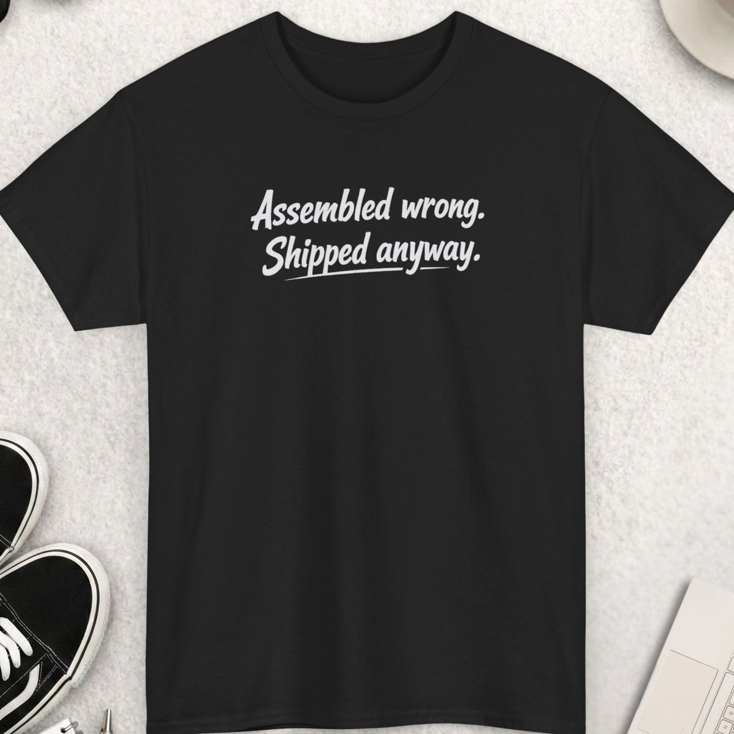 Black t-shirt with text 'Assembled wrong. Shipped anyway.' on a light gray surface with shoes, coffee, and accessories.