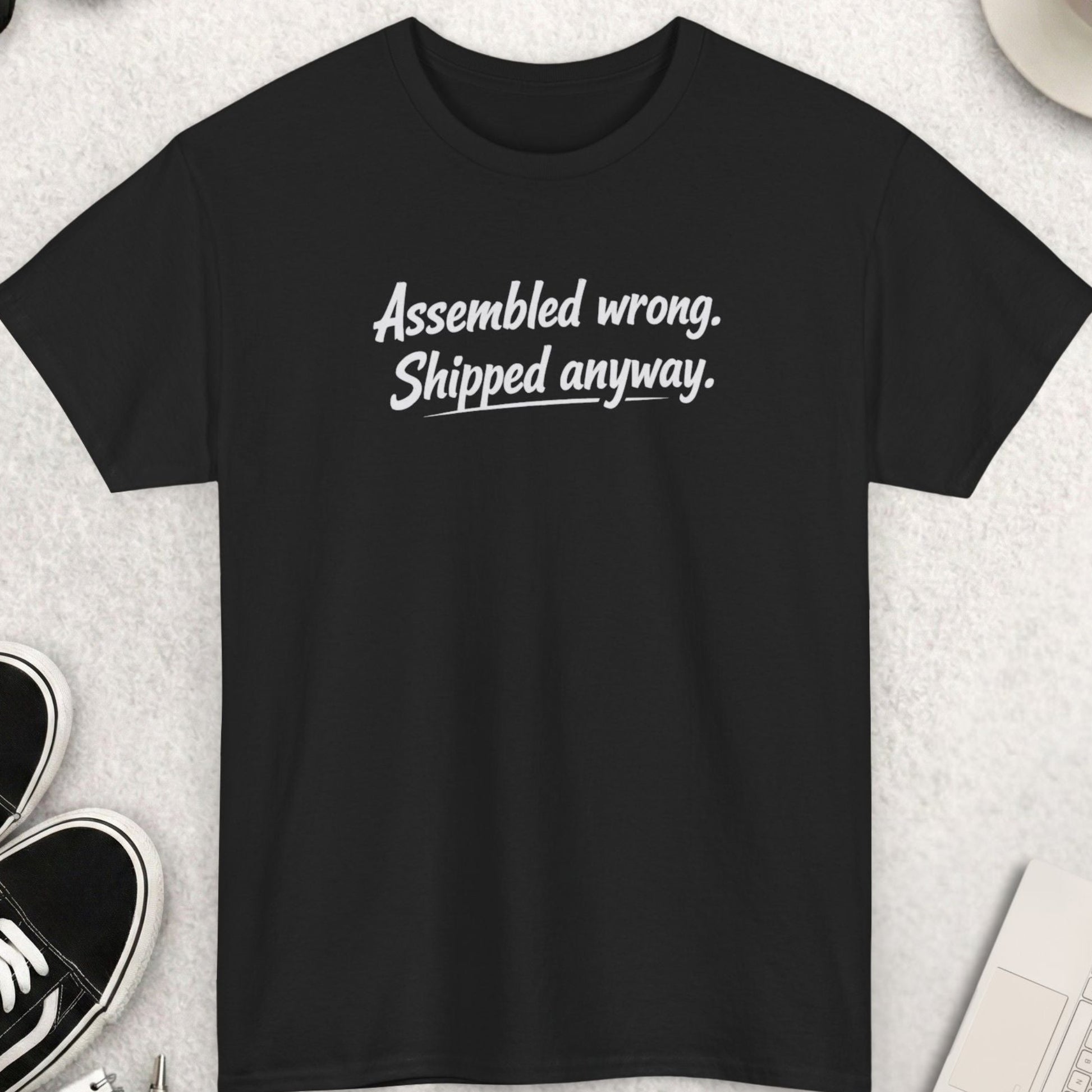 Black t-shirt with text 'Assembled wrong. Shipped anyway.' on a light gray surface with shoes, coffee, and accessories.