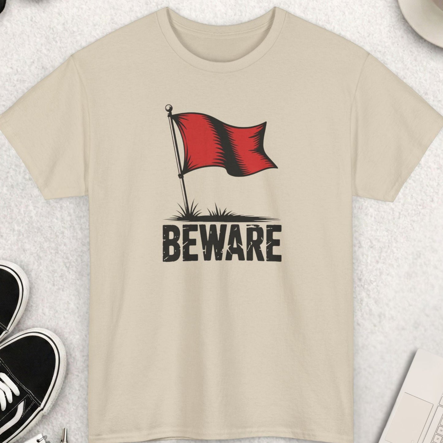 Beige t-shirt with a red flag and 'Beware' text on a light gray surface with shoes, sunglasses, and a cup of coffee.