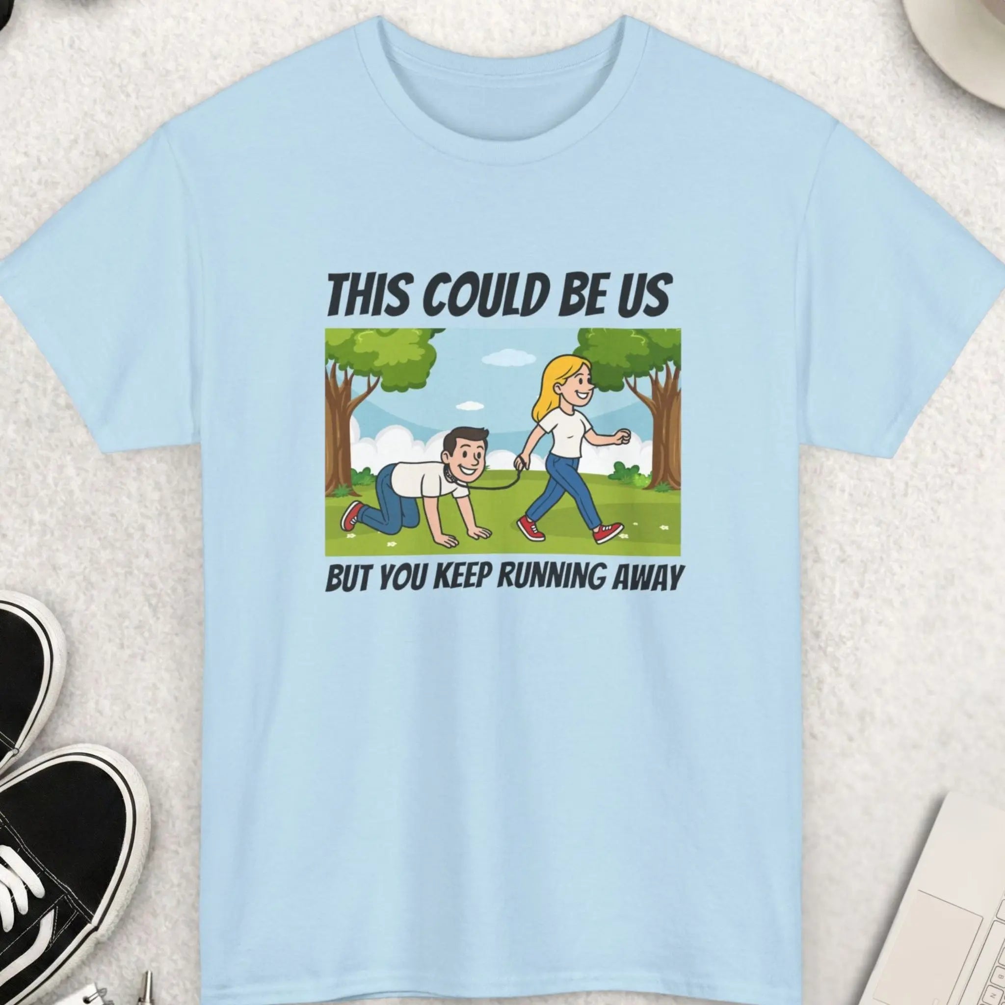 Light blue t-shirt with a graphic and text on a light gray surface with shoes, sunglasses, and a cup of coffee.