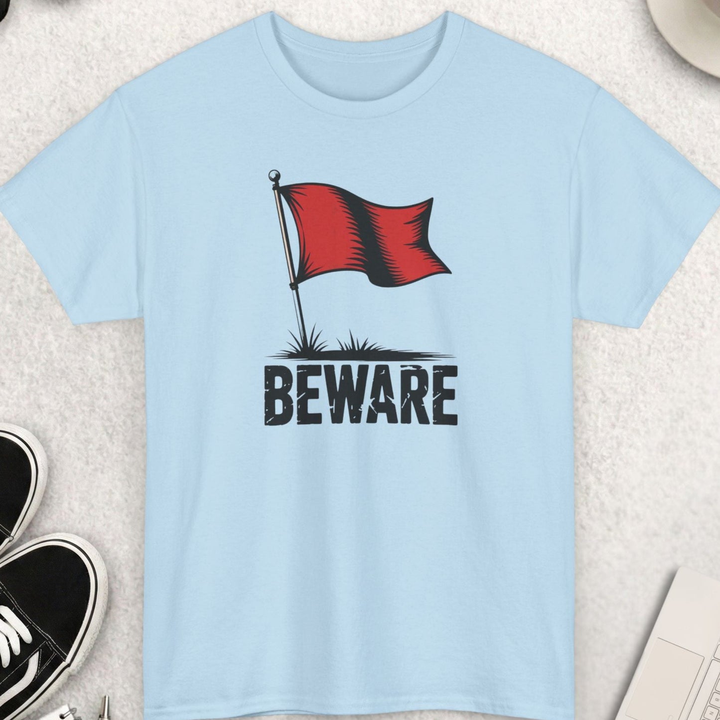 Light blue t-shirt with a red flag and 'BEWARE' text on a light gray surface with shoes, sunglasses, and a cup of coffee.