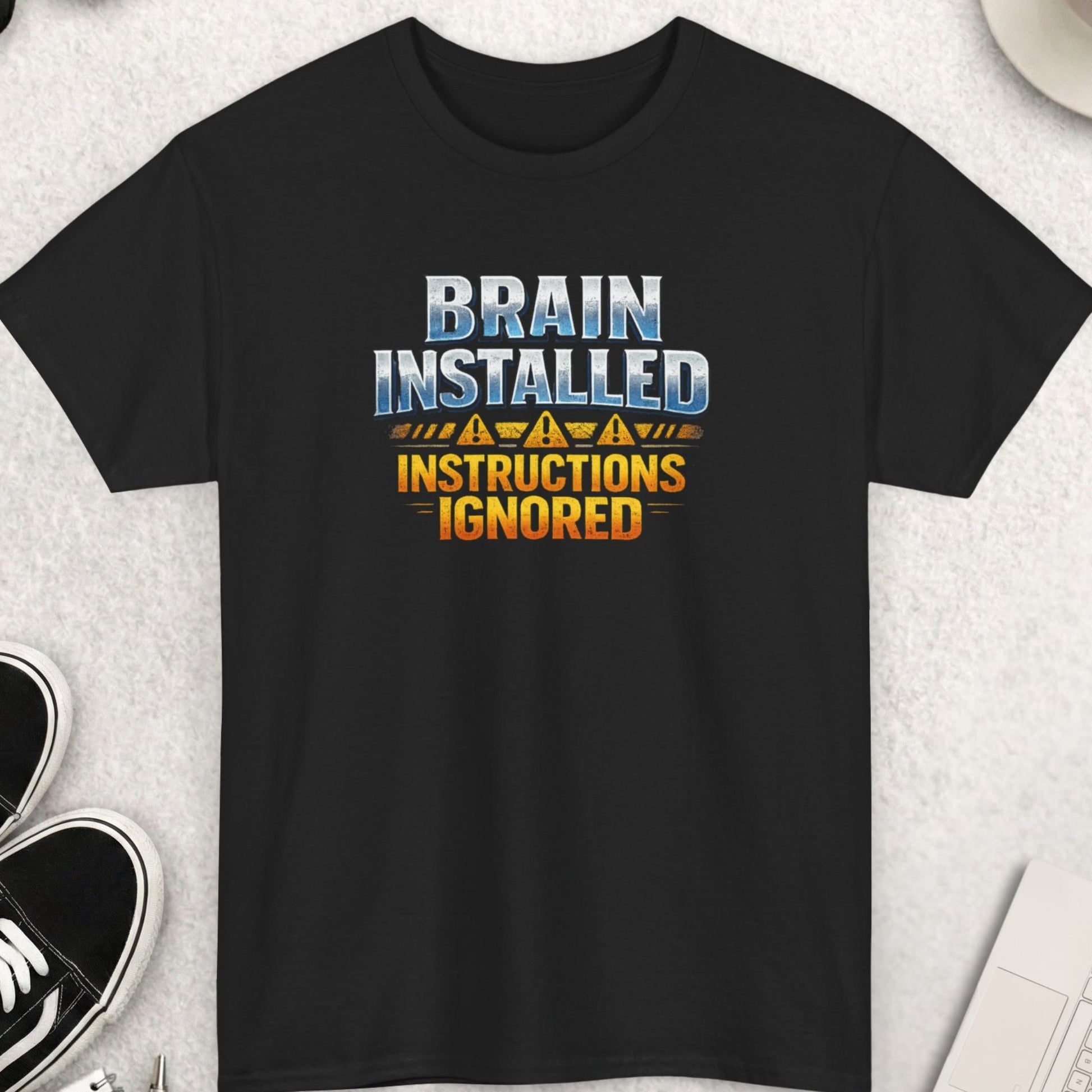 Black t-shirt with 'Brain Installed Instructions Ignored' text on a light gray surface with shoes, coffee, and accessories.