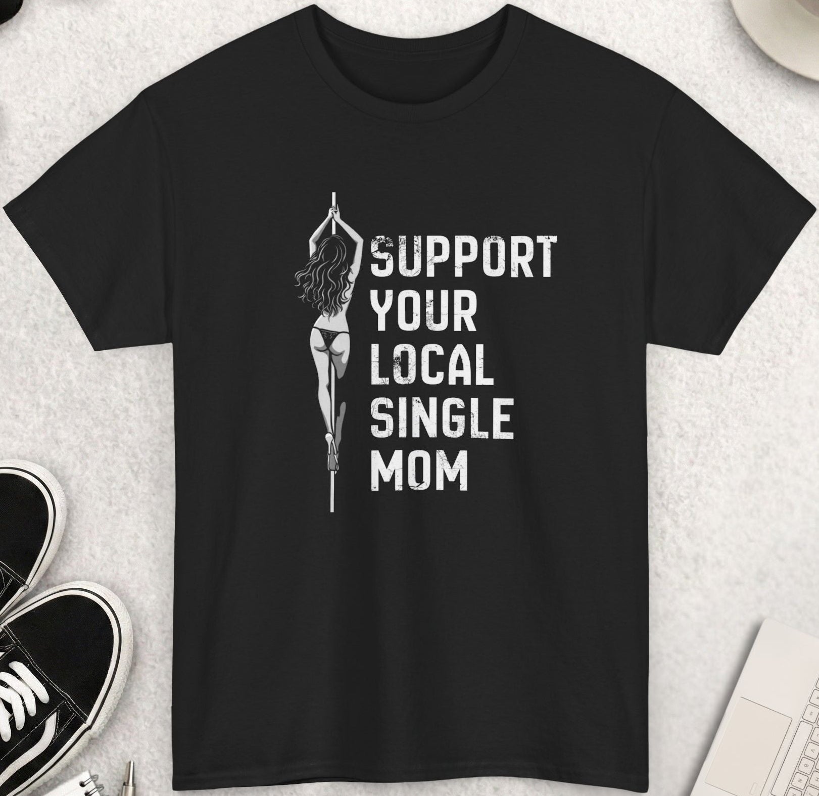 Black t-shirt with 'Support Your Local Single Mom' text on a light gray surface with shoes, coffee, and sunglasses.