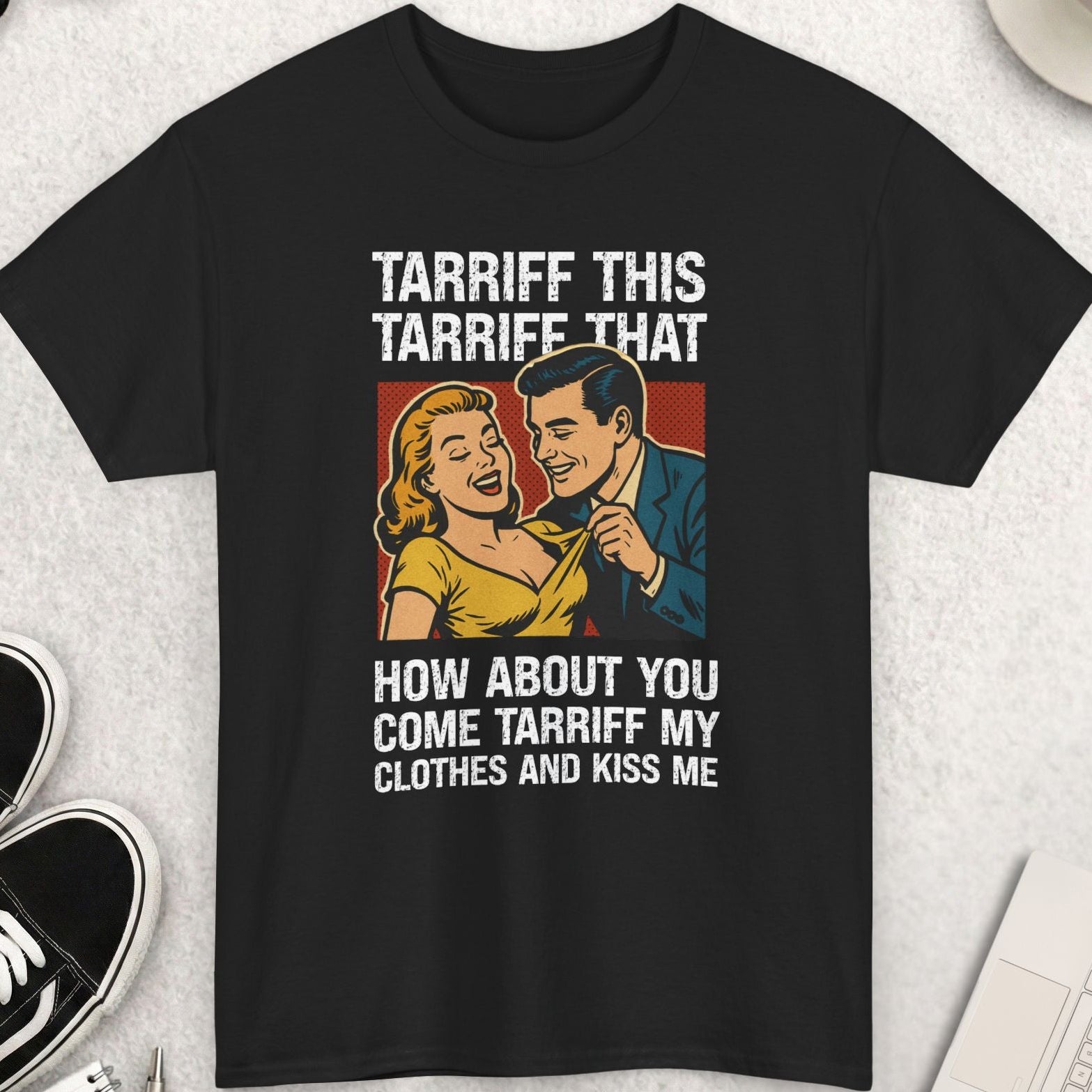 Black t-shirt with graphic design and text on a light gray surface with shoes, coffee, and sunglasses.