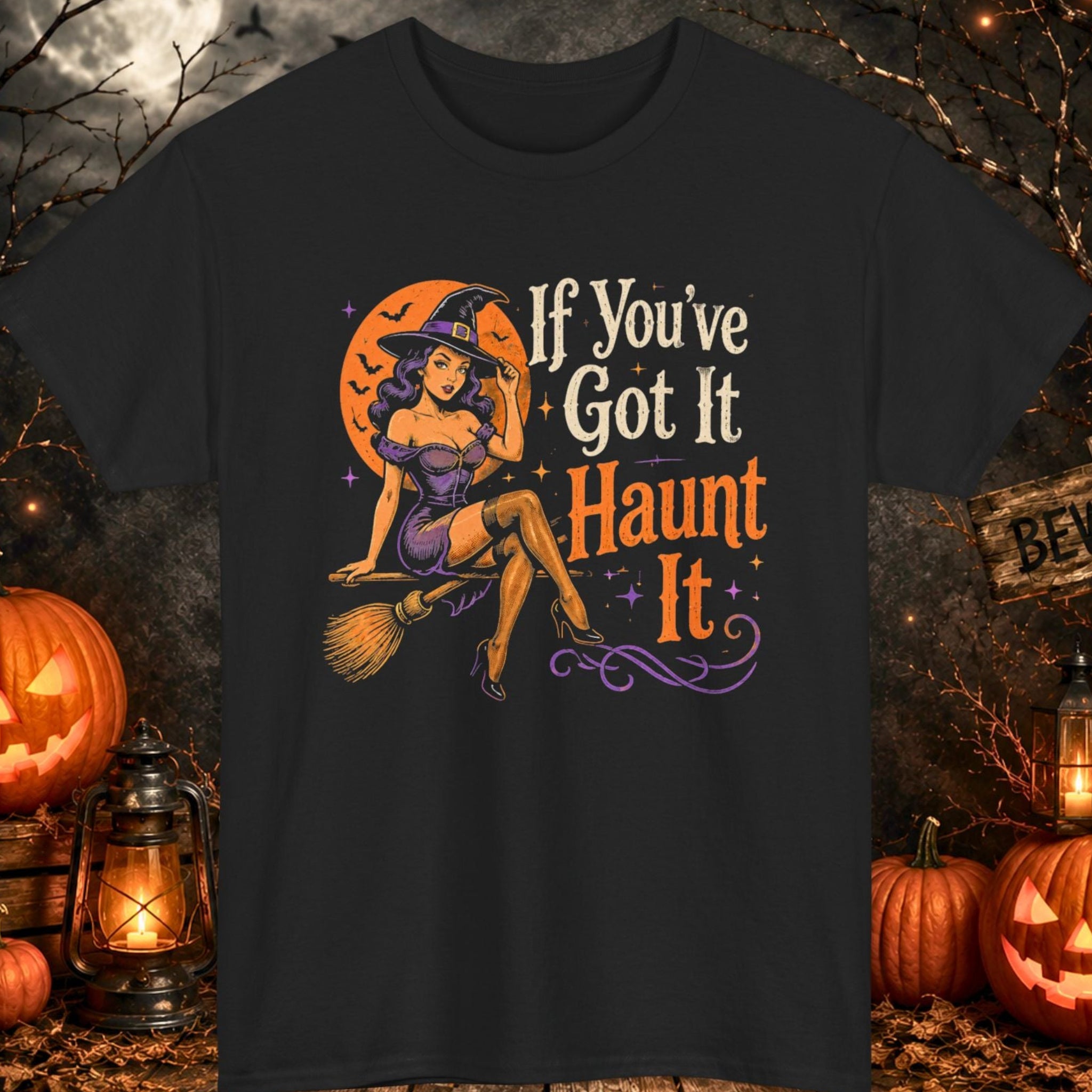 Mayhem Haus black Halloween T-shirt featuring a pin-up-inspired witch sitting on a broomstick beneath an orange moon beside the phrase "If You've Got It Haunt It." The design incorporates distressed typography, vintage Halloween artwork, purple accents, and retro spooky-season aesthetics.