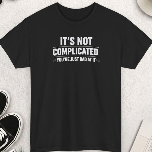 Black t-shirt featuring the sarcastic statement "IT'S NOT COMPLICATED YOU'RE JUST BAD AT IT" in bold distressed white typography. The design uses oversized vintage-style lettering with subtle weathered textures and clean divider lines, creating a humorous, blunt, and unapologetically direct aesthetic.