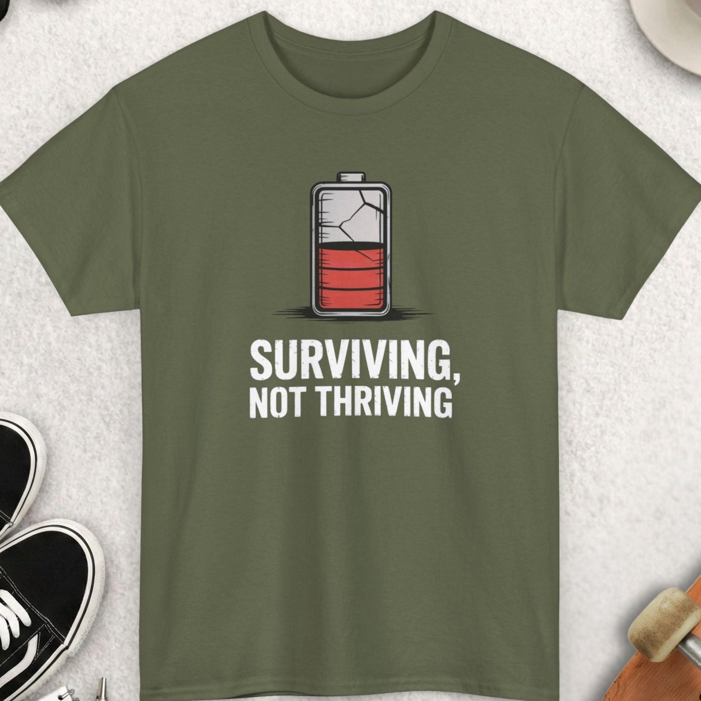 Green t-shirt with battery graphic and text 'Surviving, Not Thriving' on a light gray surface with accessories.