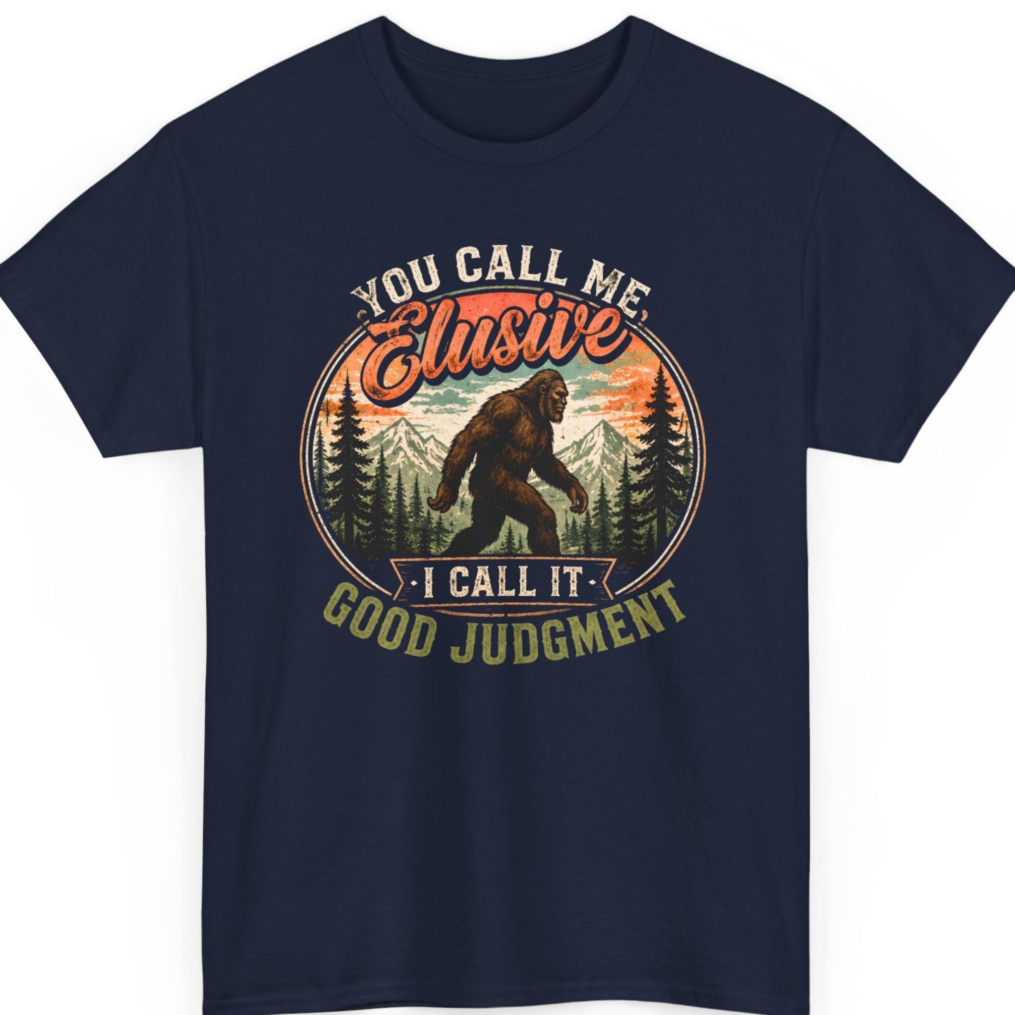Mayhem Haus Sasquatch t-shirt on a navy blue shirt featuring a classic Bigfoot silhouette walking through a forest with mountain scenery and the humorous quote "You Call Me Elusive I Call It Good Judgment."
