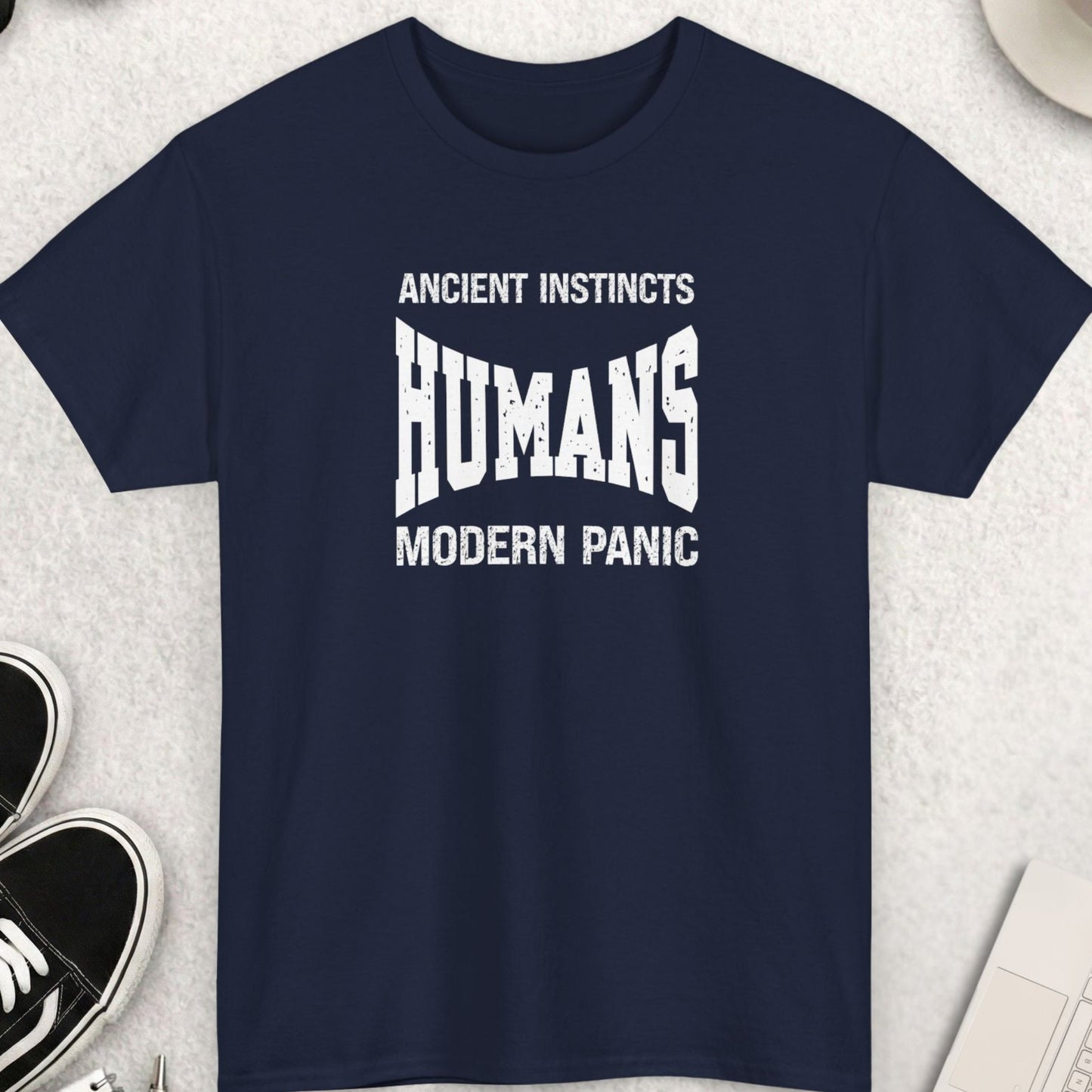 Navy blue t-shirt with 'ANCIENT INSTINCTS HUMANS MODERN PANIC' text on a light gray surface with shoes, coffee, and accessories.