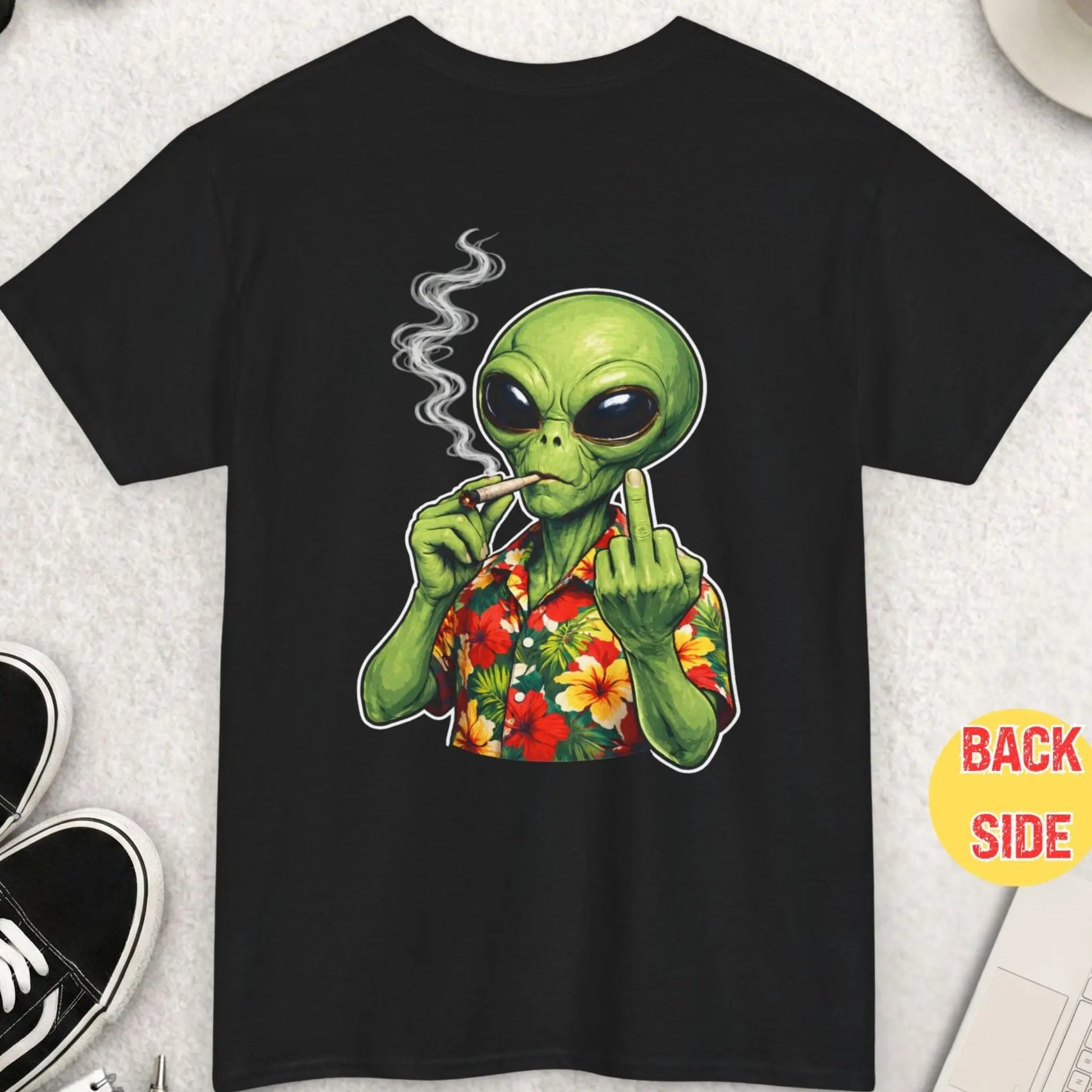 Black t-shirt with a green alien smoking a joint on a light gray surface with accessories.