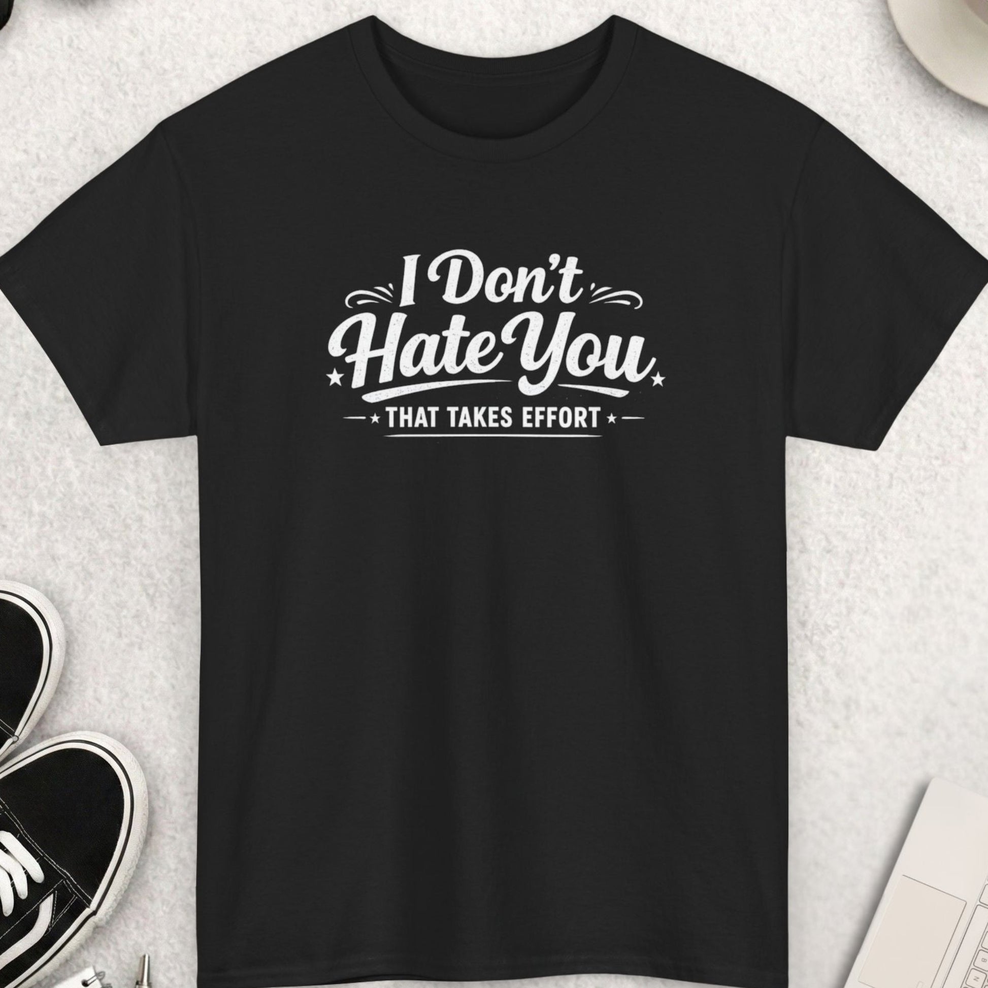 Black t-shirt with 'I Don't Hate You, That Takes Effort' text on a light gray surface with shoes, coffee, and accessories.