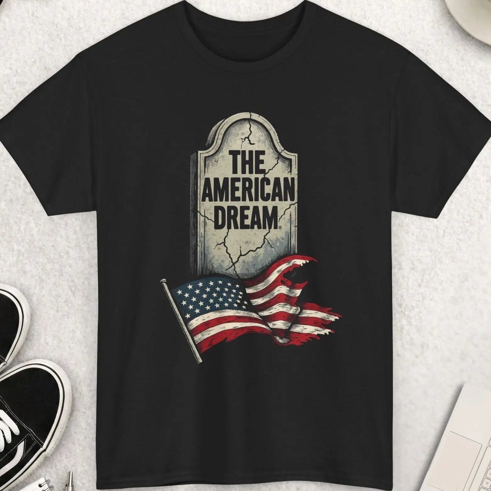 Black t-shirt with 'The American Dream' graphic on a light gray surface with shoes, coffee, and accessories.