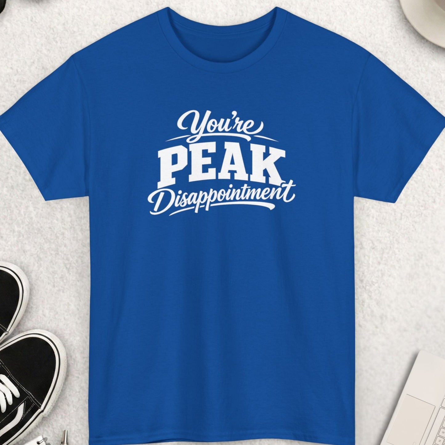 Blue t-shirt with 'You're Peak Disappointment' text on a light gray surface with accessories.