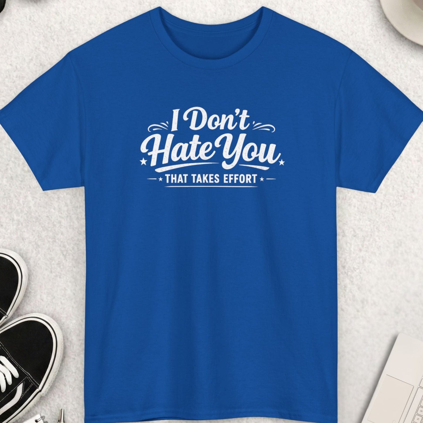 Blue t-shirt with 'I Don't Hate You That Takes Effort' text on a light gray surface with shoes, sunglasses, and coffee.