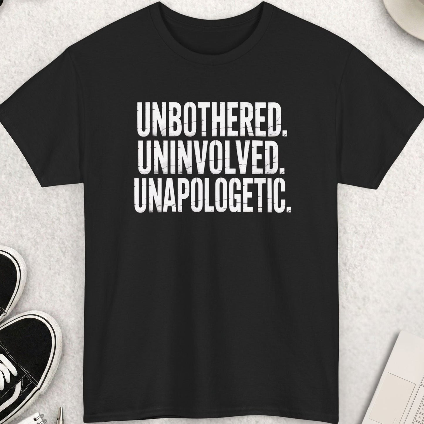 Black t-shirt with 'UNBOtherED. UNINvolved. UNApoLOGetic.' text on a light gray surface with shoes, coffee, and accessories.