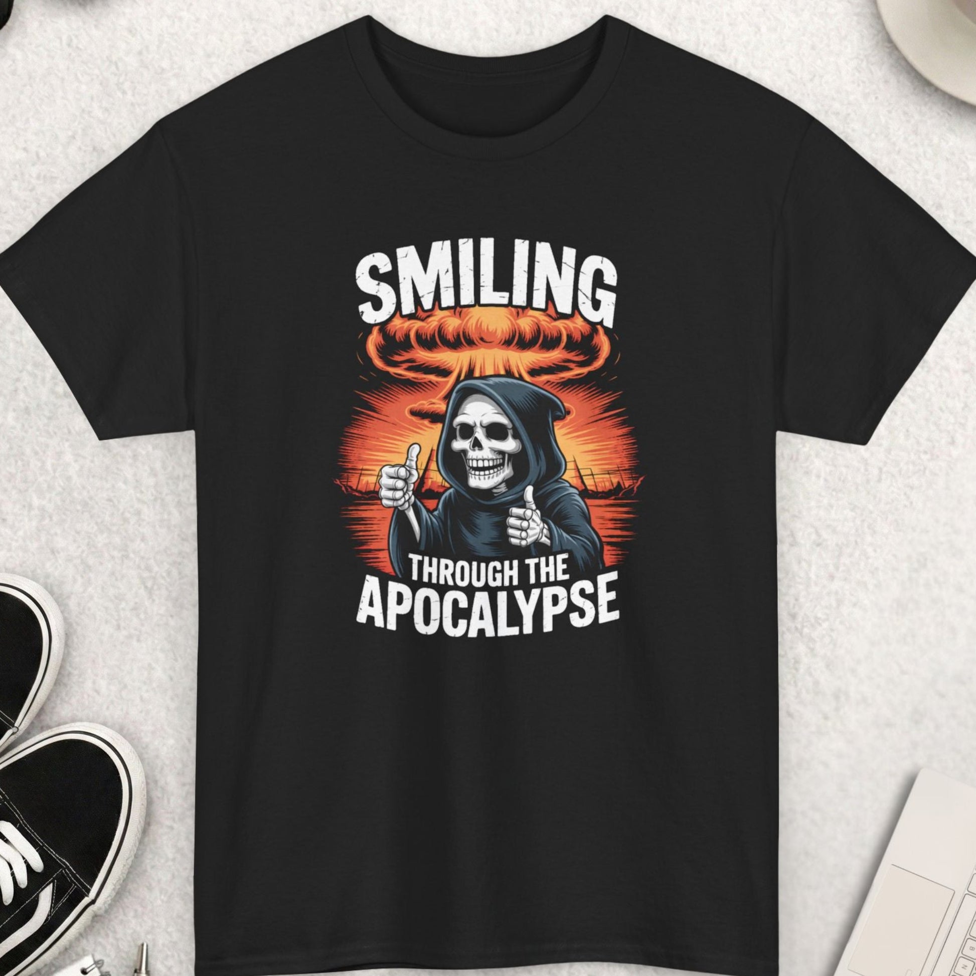 Black t-shirt with a graphic design featuring a skeleton and text 'Smiling Through the Apocalypse' on a light gray surface with shoes, sunglasses, and a cup of coffee.