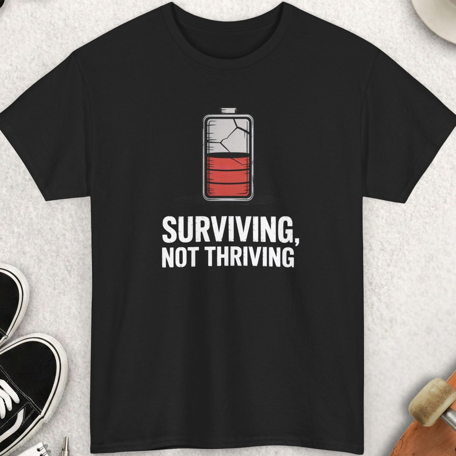 Black t-shirt with battery graphic and text 'Surviving, Not Thriving' on a light gray surface with accessories.