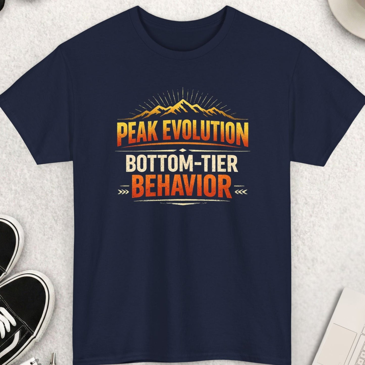 Navy blue t-shirt with 'Peak Evolution Bottom-Tier Behavior' design on a light gray surface with accessories.