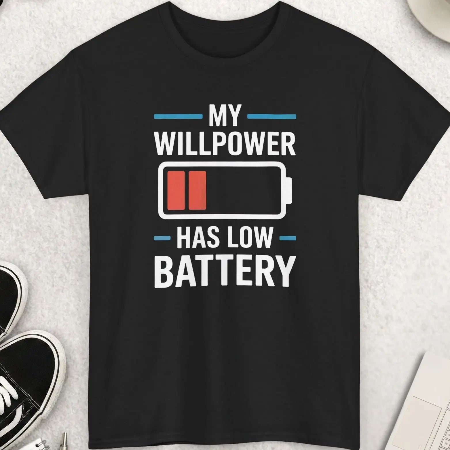 Black t-shirt with 'My willpower has low battery' design on a light gray surface with accessories.