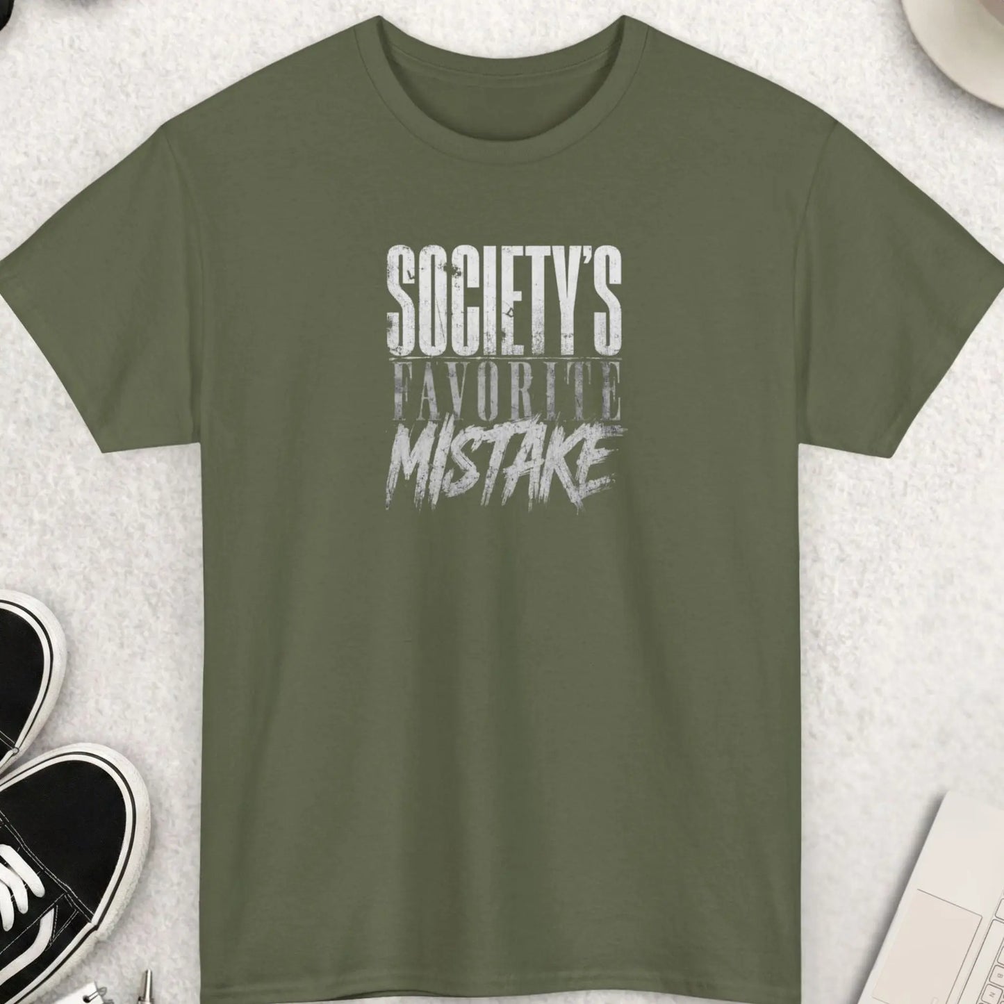 Military Green t-shirt with 'Society's Favorite Mistake' text on a light gray surface with shoes, coffee, and accessories.
