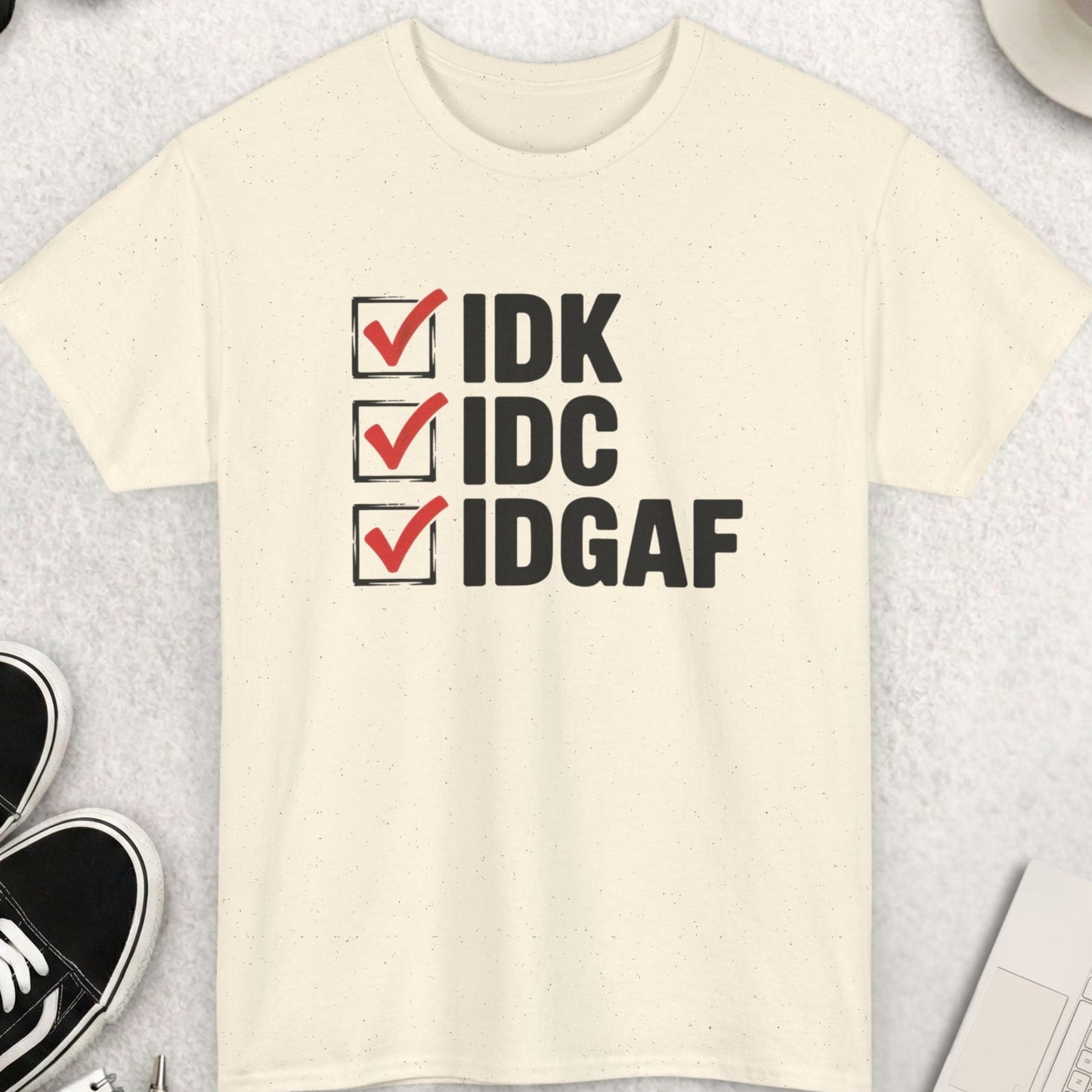 Beige t-shirt with text 'IDK IDCIDGAF' on a light gray surface with shoes, coffee, and sunglasses.