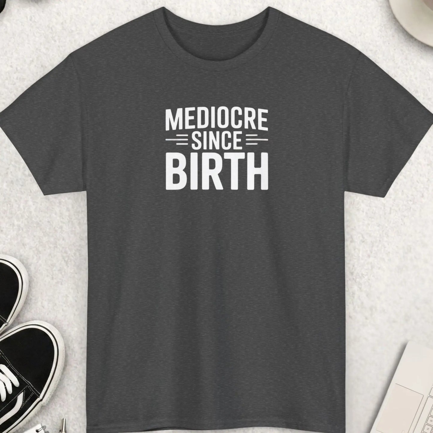Dark gray t-shirt with 'Mediocre Since Birth' text on a light gray surface with accessories.