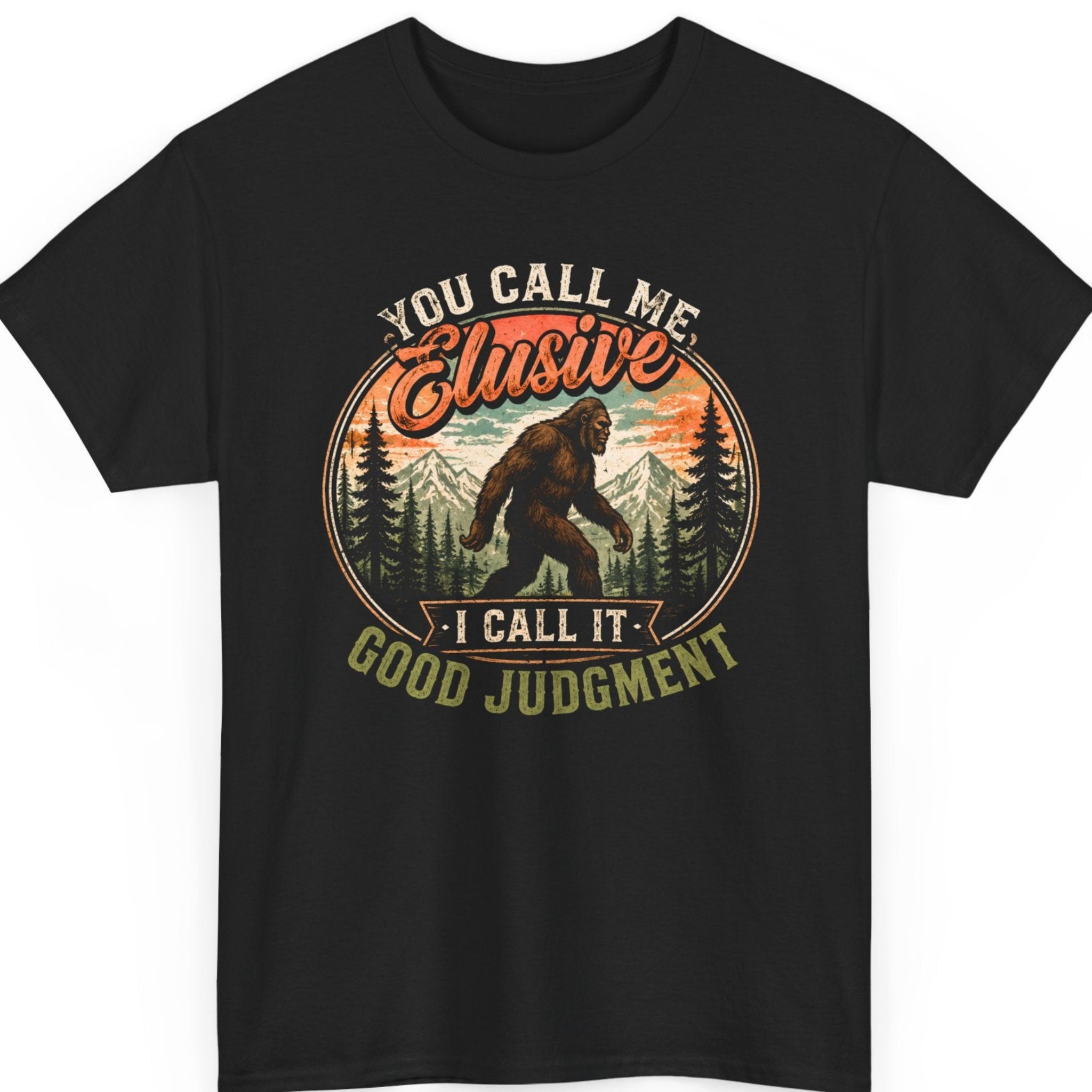 Mayhem Haus Bigfoot graphic t-shirt on a black shirt featuring a walking Sasquatch surrounded by pine trees and mountains inside a vintage circular badge with the phrase "You Call Me Elusive I Call It Good Judgment" in distressed retro typography.