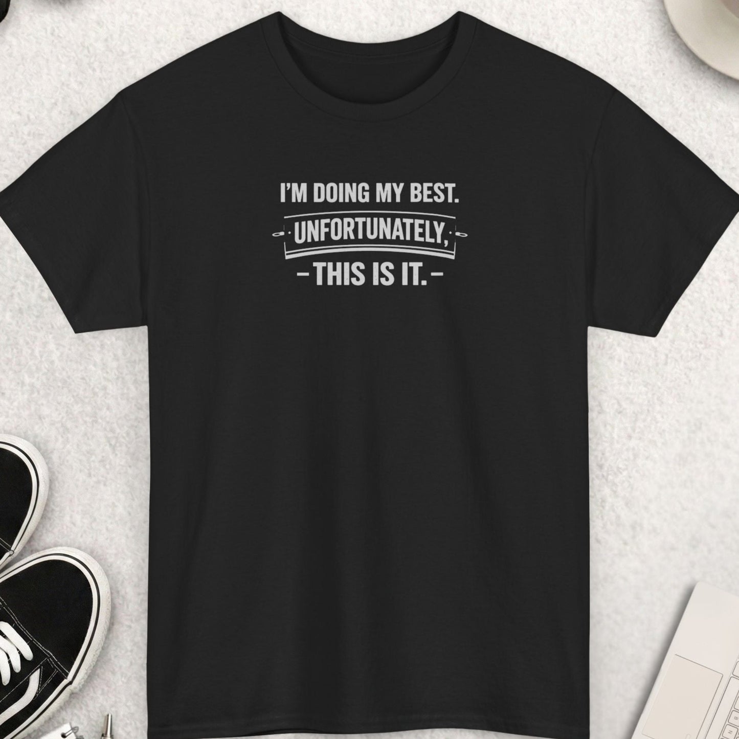 Black t-shirt with humorous text on a light gray surface with shoes, coffee, and accessories.