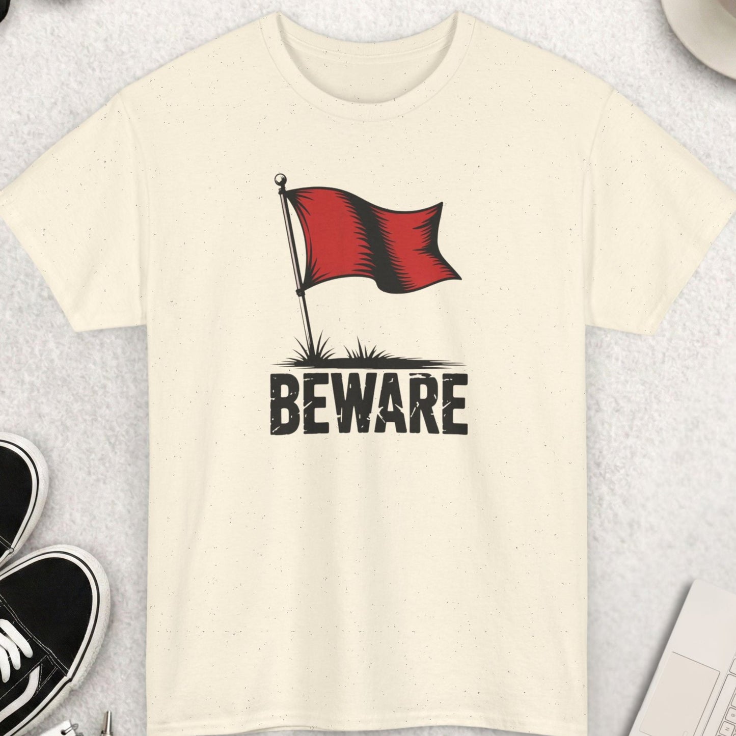 Beige t-shirt with a red flag and 'Beware' text on a light gray surface with shoes, coffee, and accessories.