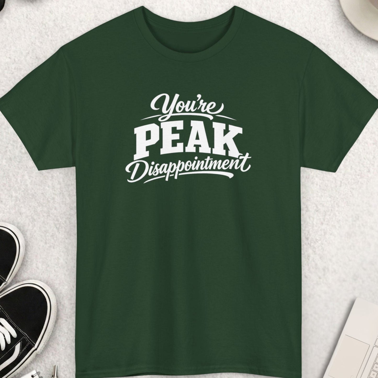 Green t-shirt with 'You're Peak Disappointment' text on a light gray surface with shoes, coffee, and sunglasses.