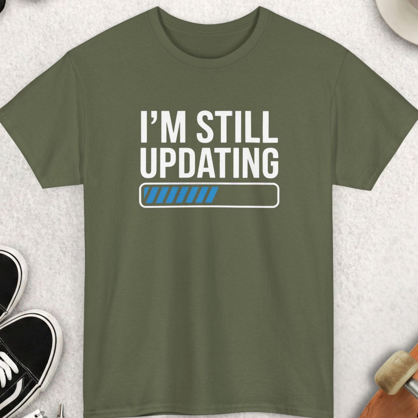 Green t-shirt with 'I'm Still Updating' text on a light gray surface with accessories.