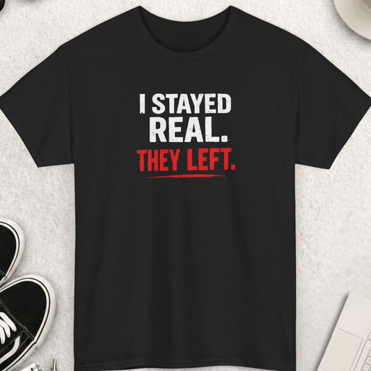 Black t-shirt with 'I Stayed Real. They Left.' text on a light gray surface with shoes, coffee, and accessories.