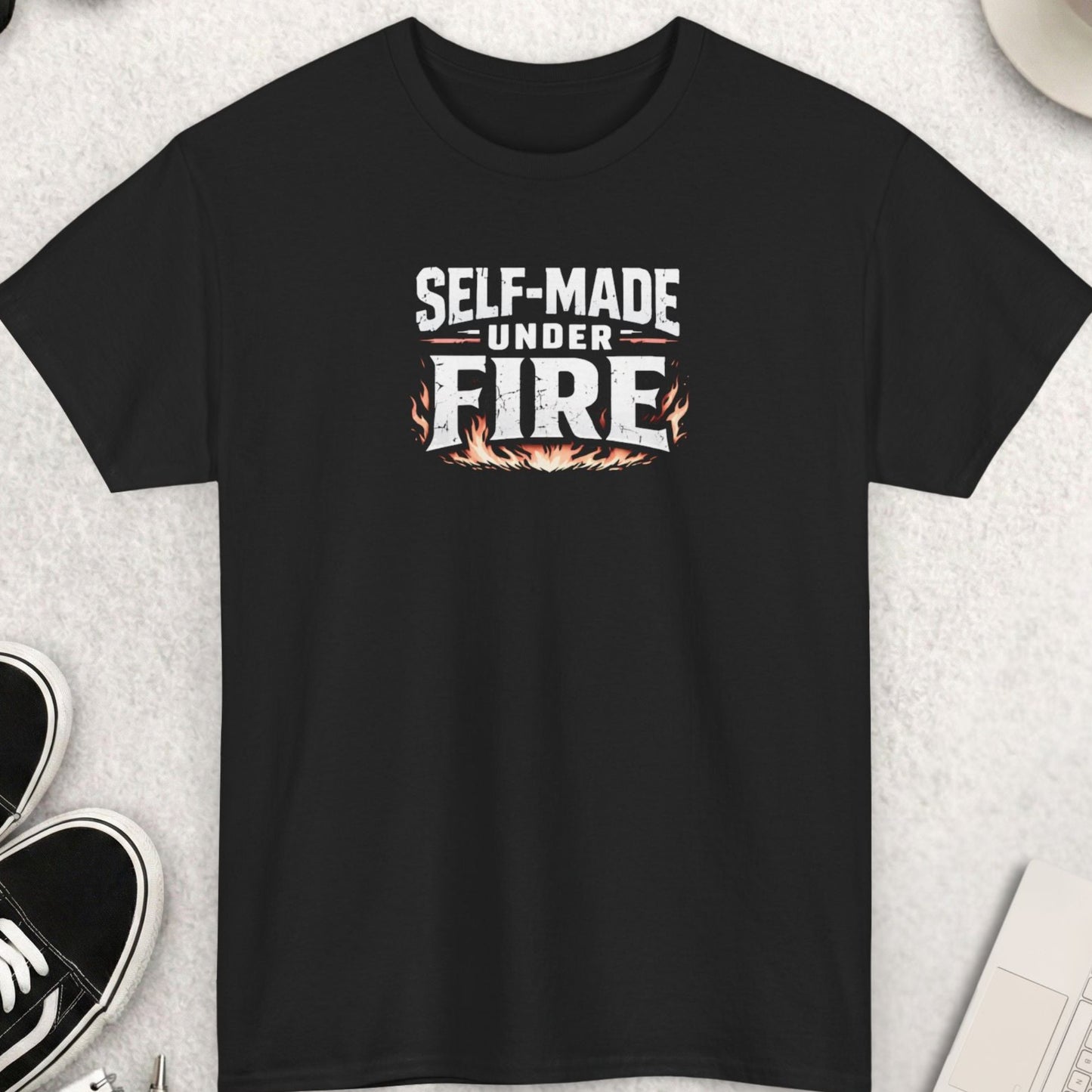 Black t-shirt with 'Self-Made Under Fire' text on a light gray surface with shoes, coffee, and sunglasses.