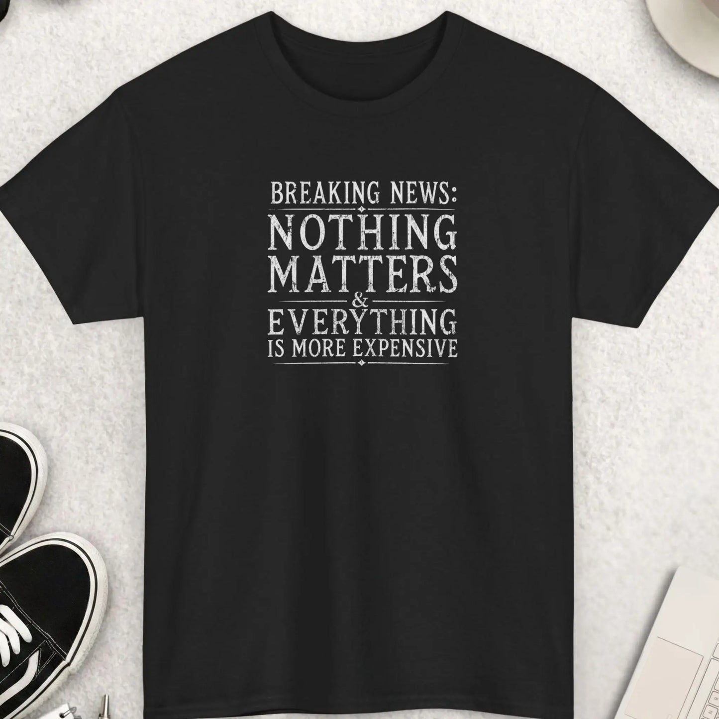 Breaking News: Nothing Matters & Everything Is More Expensive" vintage distressed newspaper-style graphic t-shirt on a black background. Bold all-white serif typography with ornate diamond dividers and double-rule lines in a worn vintage newsprint aesthetic. Funny sarcastic inflation and dark humor statement tee.