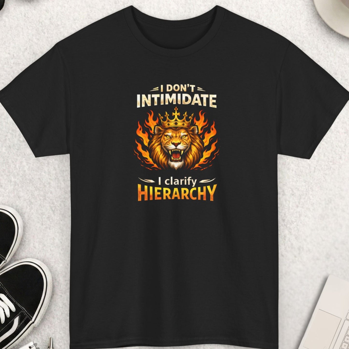 Black t-shirt ' I Don’t Intimidate – I Clarify Hierarchy' on a light gray surface with shoes, coffee, and accessories.