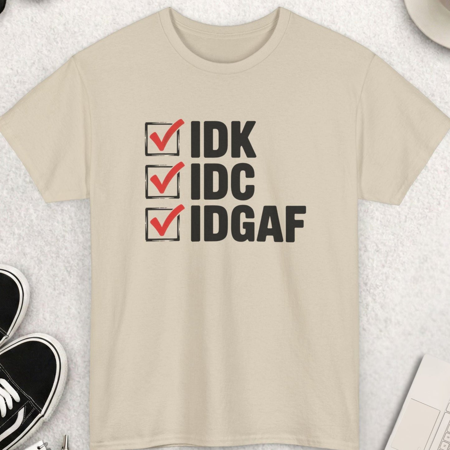 Beige t-shirt with text 'IDK', 'IDC', 'IDGAF' on a light gray surface with shoes, sunglasses, and a cup of coffee.